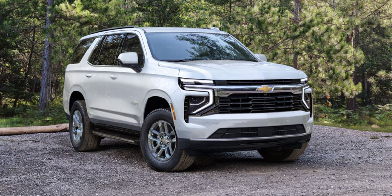 White Chevrolet Tahoe SUV parked outdoors on a gravel road surrounded by green pine trees, showcasing its bold front grille and modern headlights in a natural forest setting.