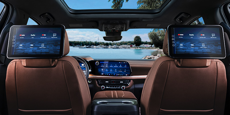 Rear-seat view of a Chevrolet Suburban modern SUV interior featuring two headrest-mounted entertainment screens and a large central dashboard display, with a lake and trees visible through the windows.